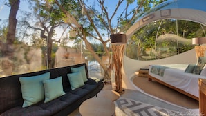 A modern, open-plan living area with a sofa, a small round table, and a bed with a view of a pool and trees. - Bubble Lodge Ile aux Cerfs (Trou d'Eau Douce)