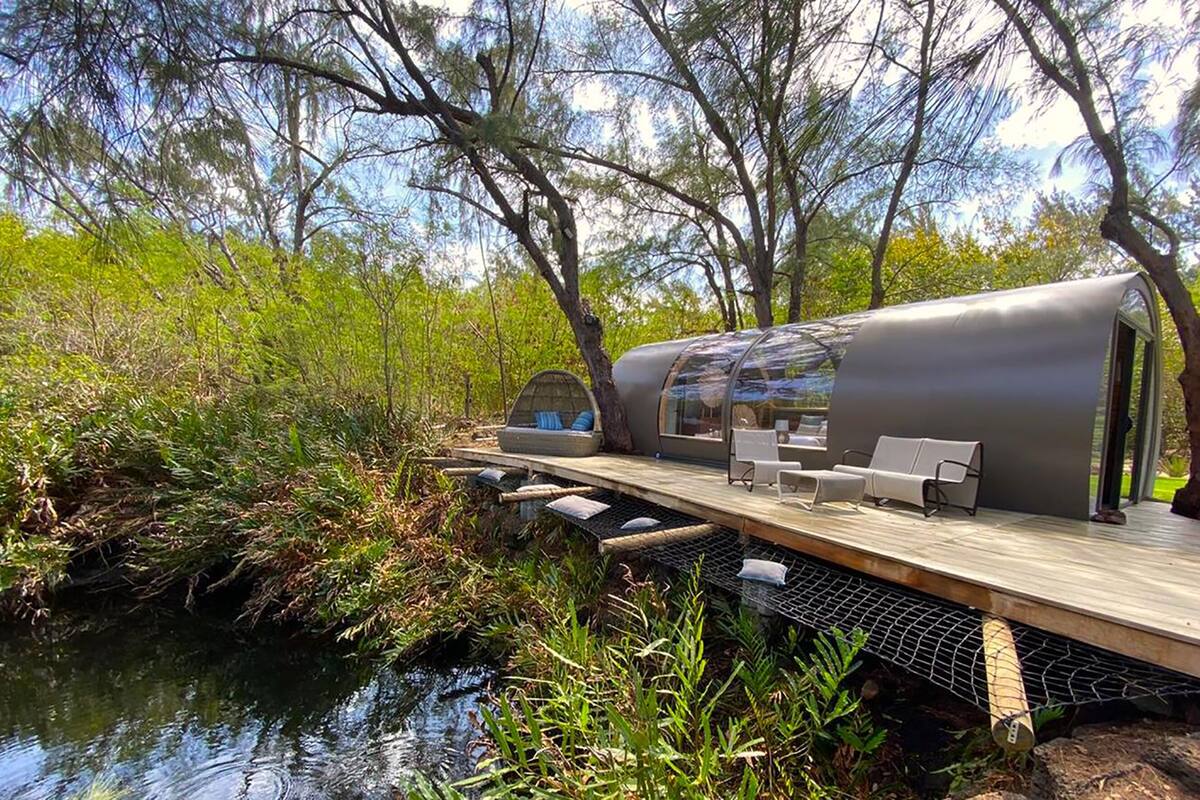 A modern, cylindrical, glass-enclosed structure with a deck and seating area, surrounded by natural vegetation and a water body.