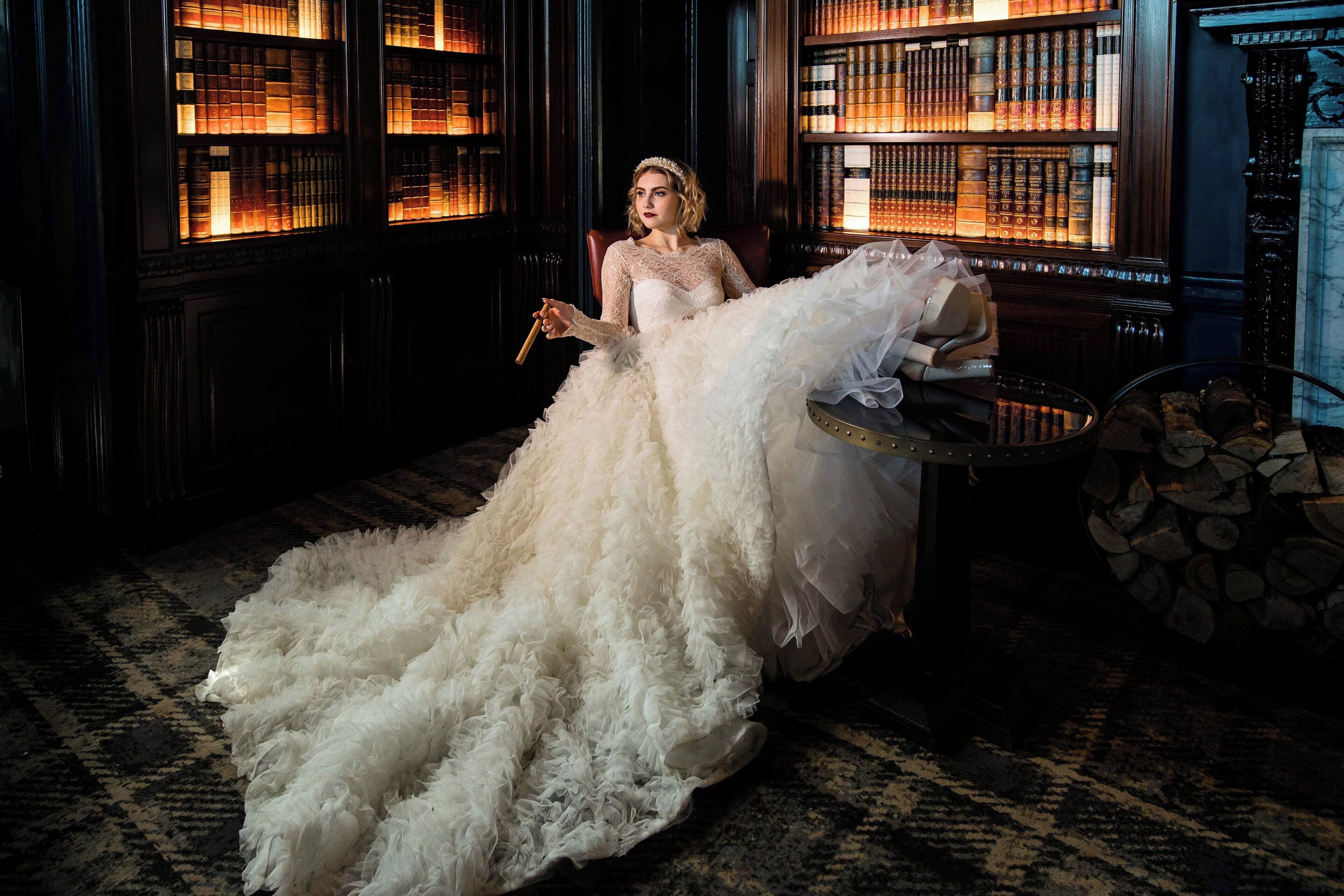 A person in a white, feathered gown seated in a room with bookshelves and a fireplace.