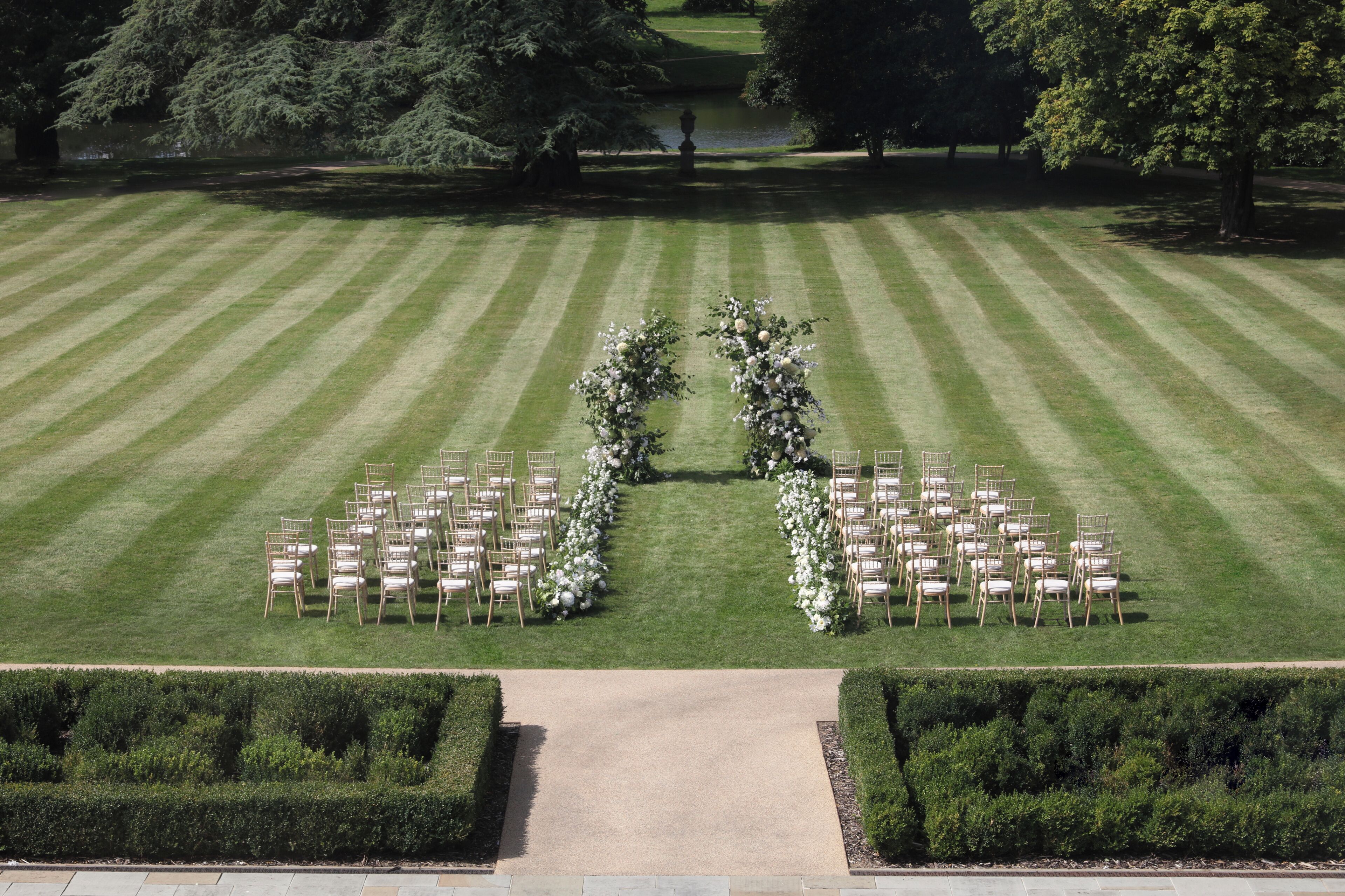 Une vue aérienne d’une pelouse agrémentée d’une arche fleurie et de rangées de chaises, probablement pour un événement.
