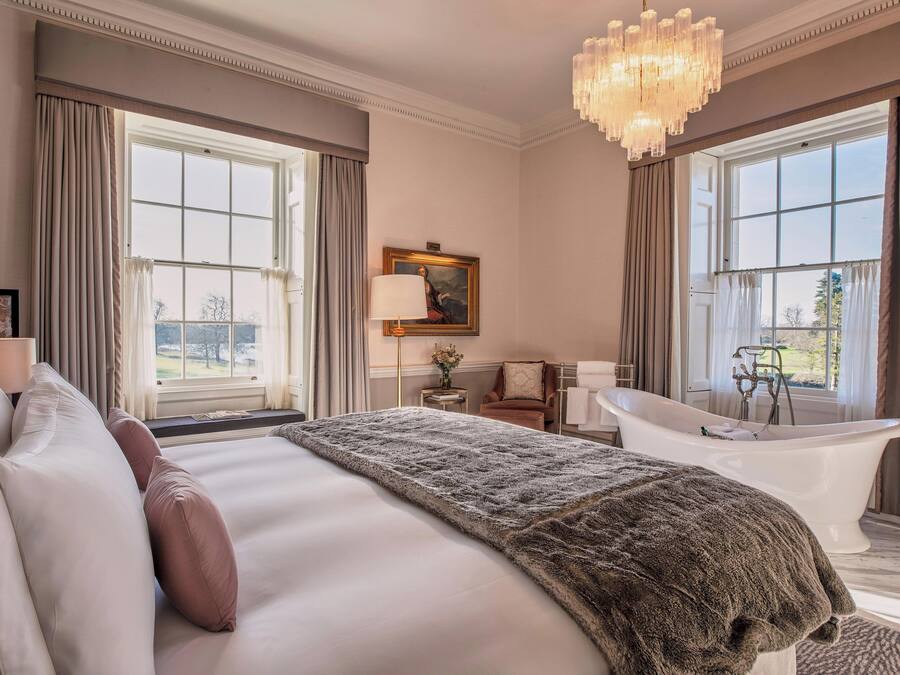 A spacious bedroom with a large bed, a bathtub, and a view of the outdoors through large windows.