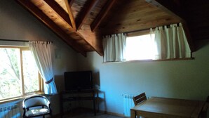 Una stanza con soffitto spiovente, una televisione su un supporto, un tavolo da pranzo e una finestra con tende.