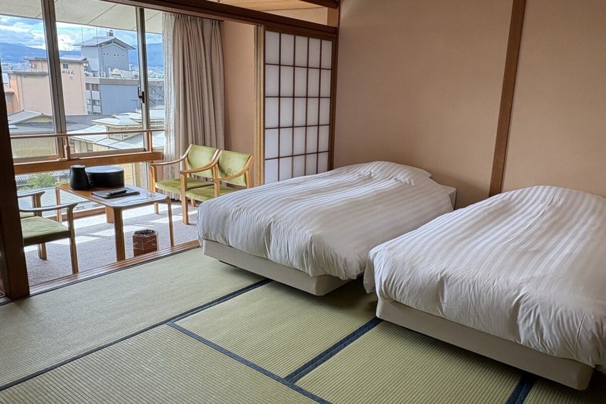 A hotel room with two beds, a tatami mat floor, a view of buildings, and a small table with a chair.