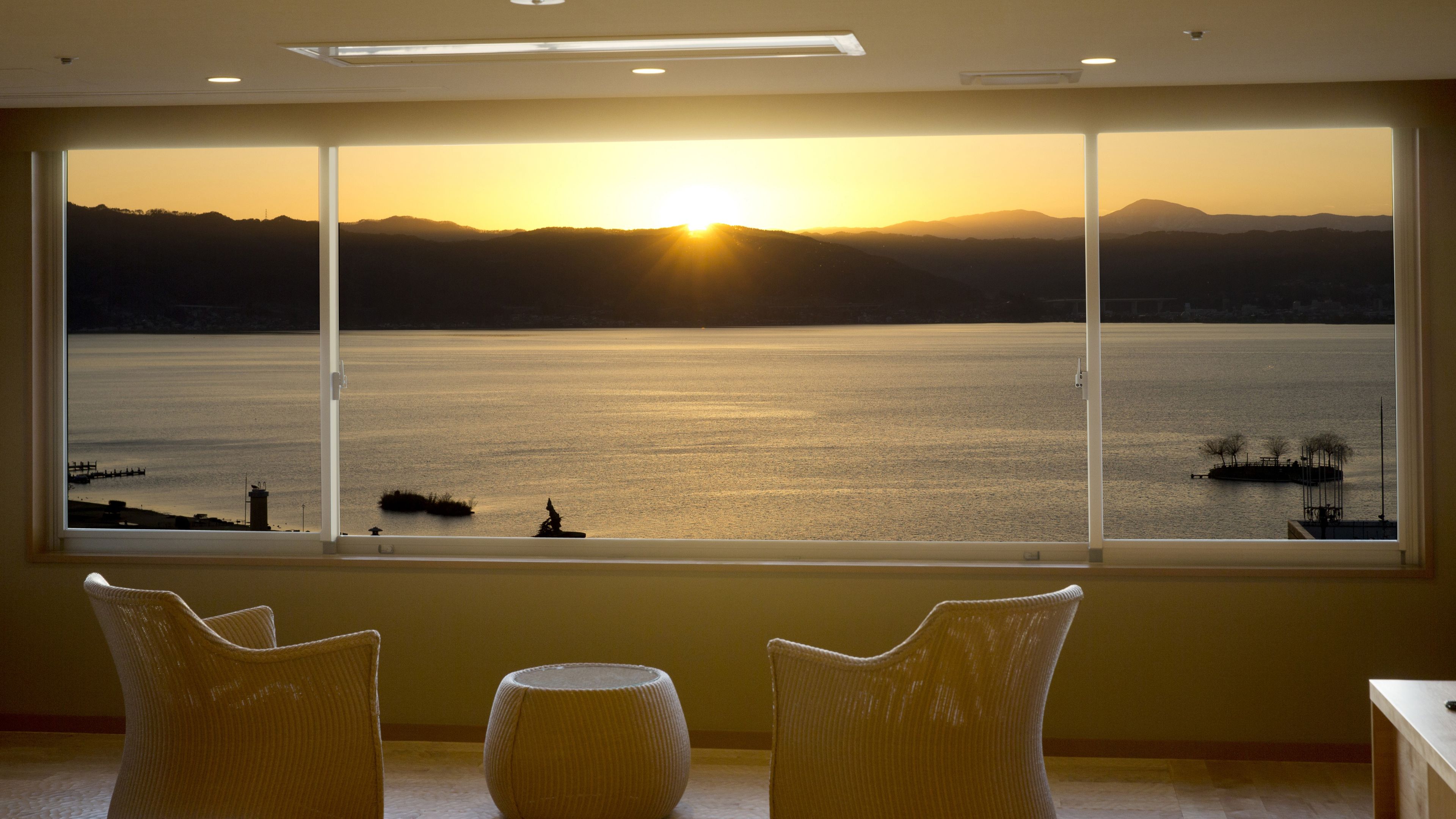 Una habitación con un amplio ventanal que da a una masa de agua y montañas distantes al atardecer.