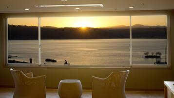 Una habitación con un amplio ventanal que da a una masa de agua y montañas distantes al atardecer.