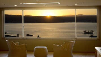 Una habitación con un amplio ventanal que da a una masa de agua y montañas distantes al atardecer.