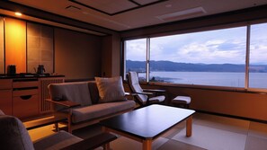 A modern interior with a sofa, chairs, a table, and a large window with a view of a body of water and mountains. - Hamanoyu (Suwa)
