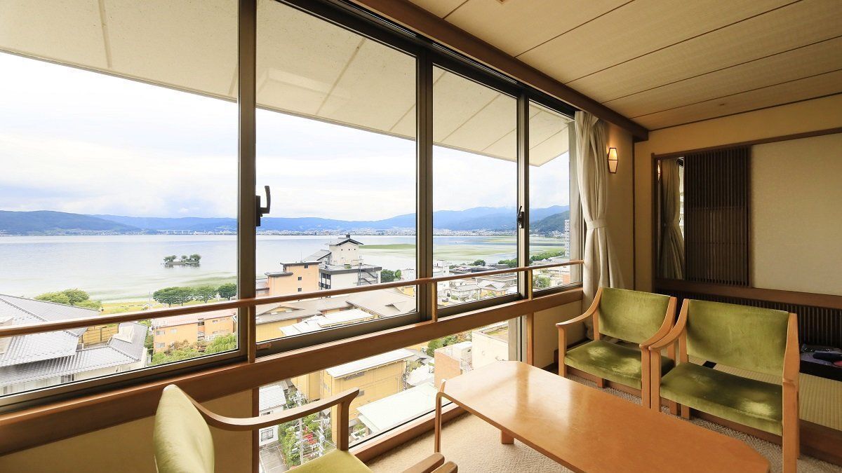 Una habitación con un ventanal grande, una mesa de madera, dos sillas verdes y vistas a un cuerpo de agua y edificios.