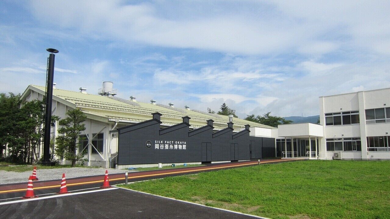 A modern building with a flat roof, a sign reading 'SILK FACT OHAYA,' and a grassy area in front.