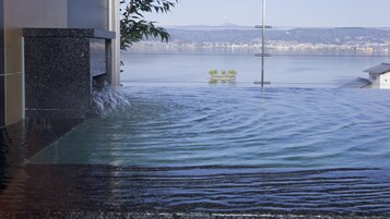 Área alagada com água corrente sobre uma estrutura, uma ponte ao fundo e vista de um horizonte urbano distante.