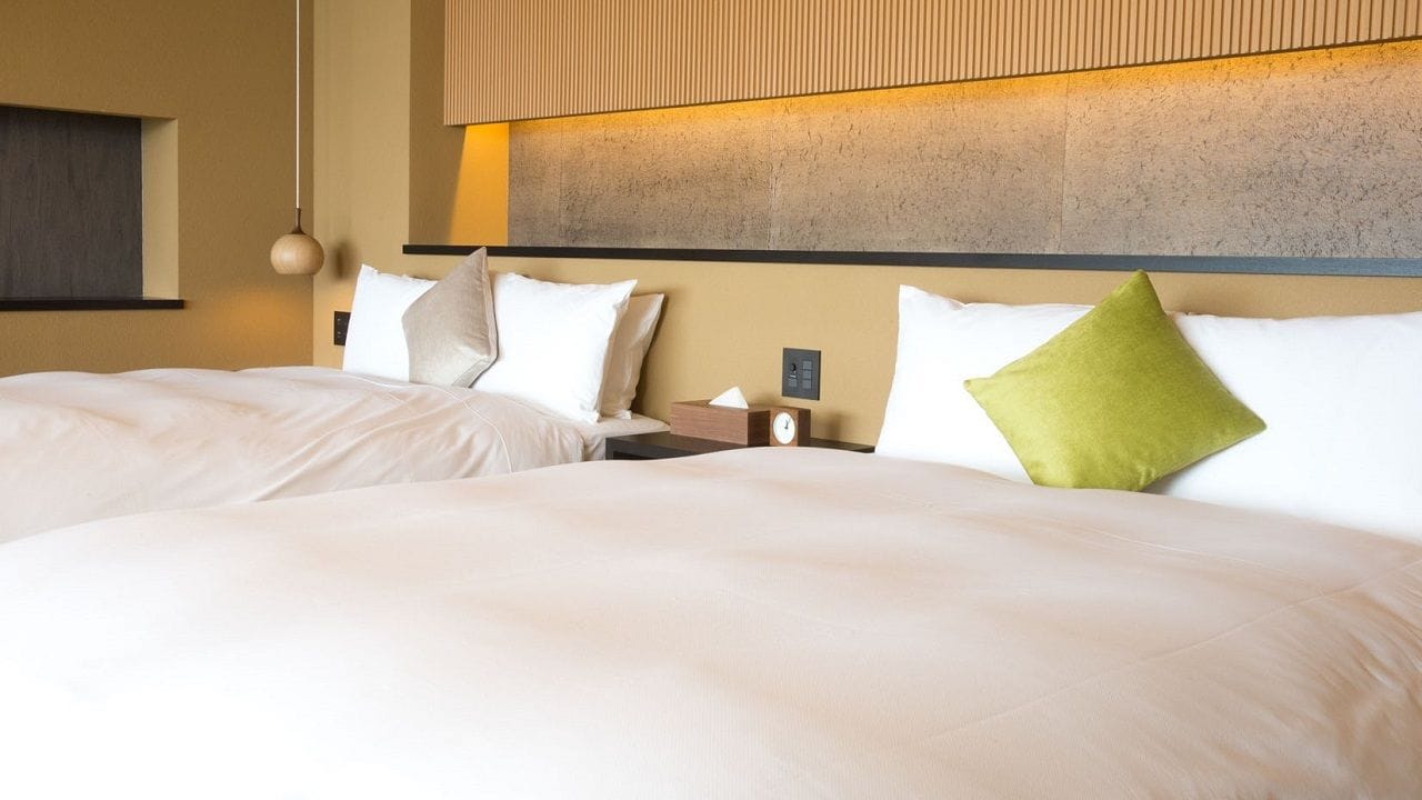 A hotel room with two beds, a nightstand with a clock, and a wall-mounted light fixture.