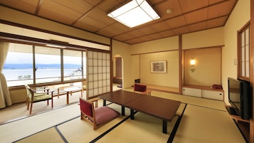 Una habitación japonesa tradicional con suelo de tatami, una mesa baja, un sofá, un televisor y vistas a un cuerpo de agua.