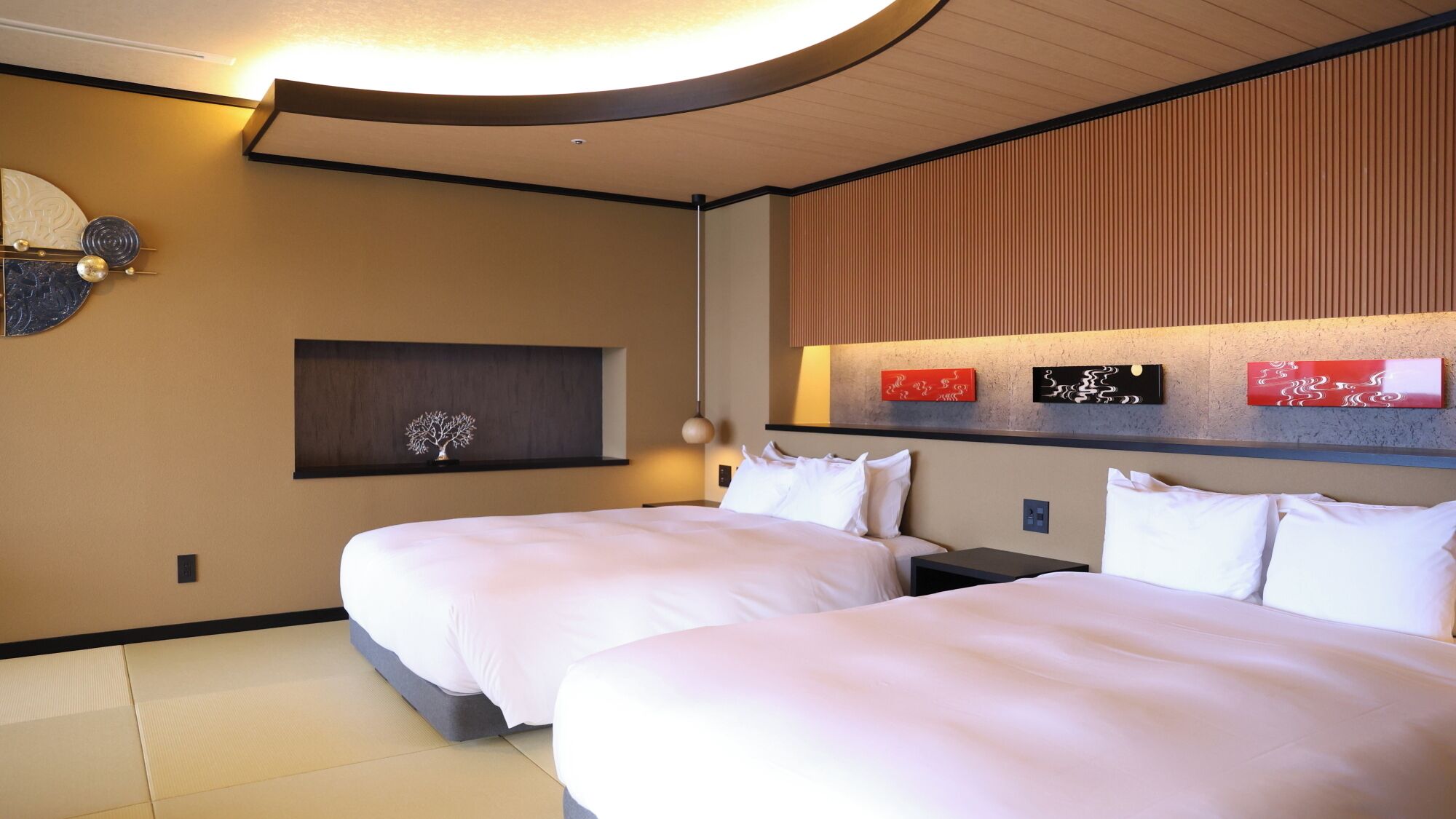 Habitación de hotel con dos camas, un televisor montado en la pared, un estante pequeño con un objeto decorativo y una lámpara en el techo.