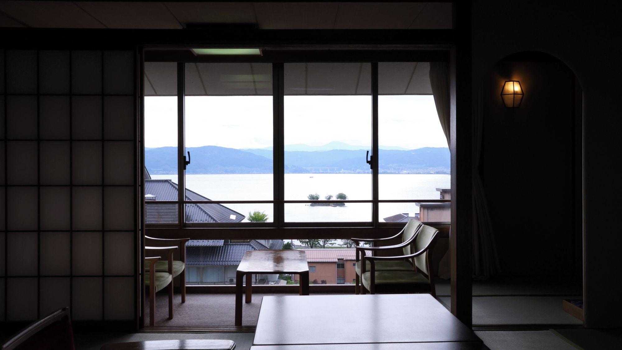 A room with a large window overlooking a body of water and mountains. There are two chairs and a table in the room.