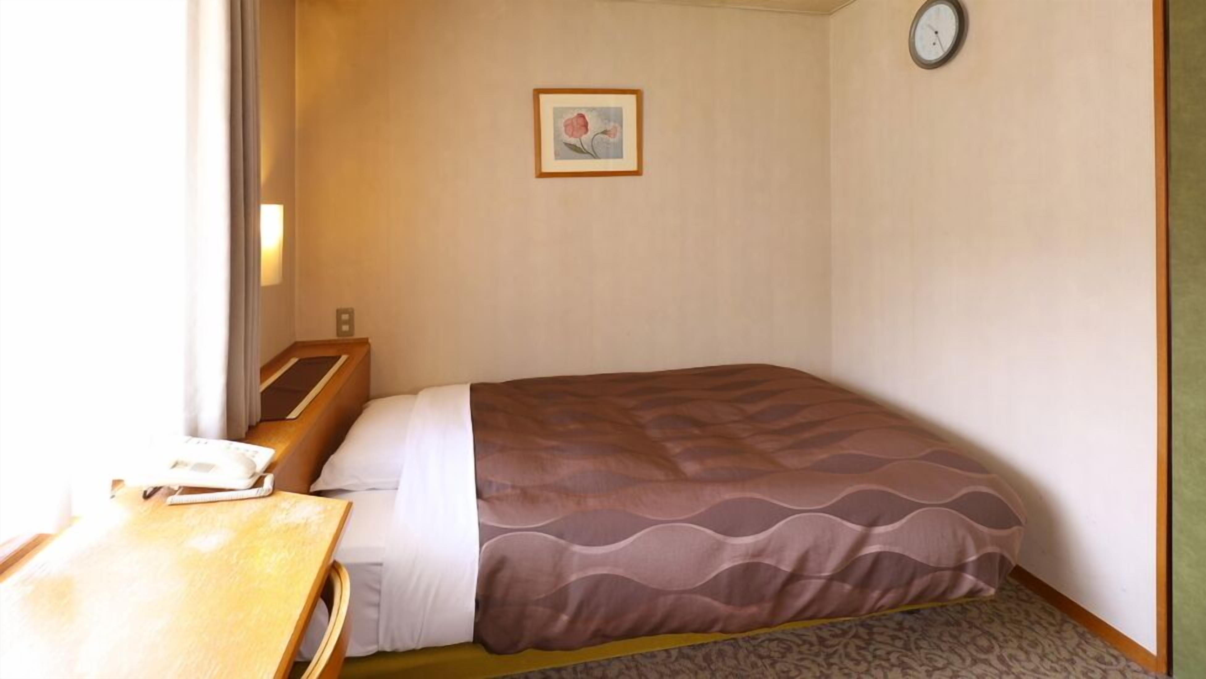Una habitación de hotel pequeña con una cama individual, un escritorio de madera, un reloj de pared y un cuadro enmarcado.