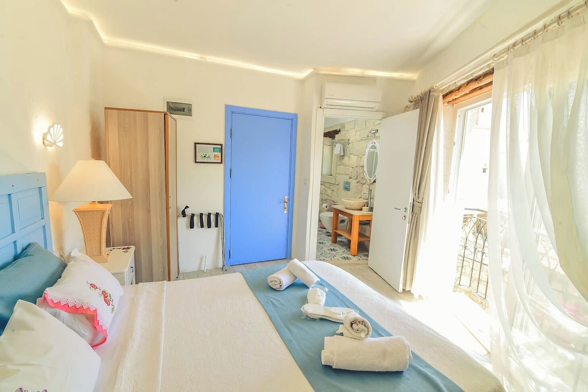 A hotel room with a bed, a blue door, a bathroom visible through an open door, and a window with curtains.