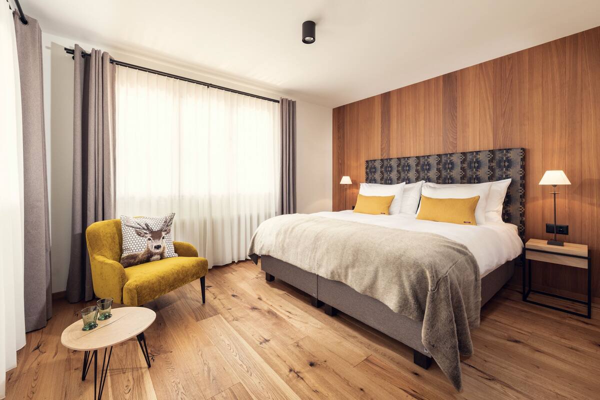 A hotel room with a large bed, a small round table, a yellow armchair, and a wooden headboard.