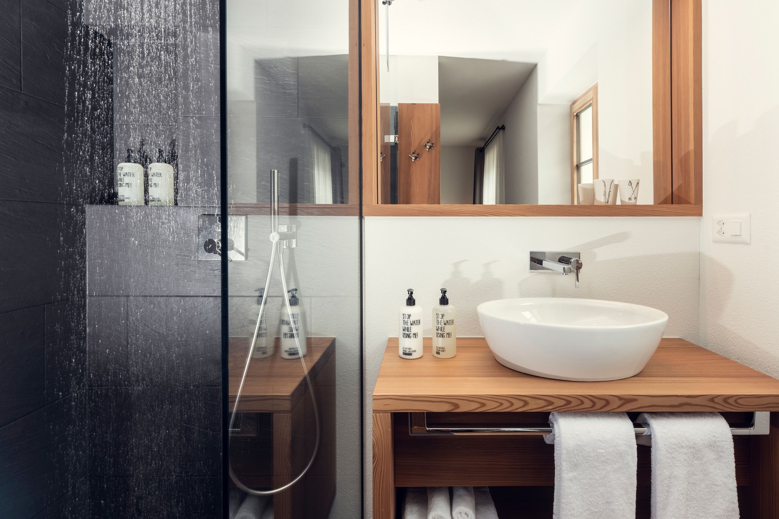 A modern bathroom with a glass-enclosed shower, a wooden vanity with a white sink, and two bottles of shampoo.