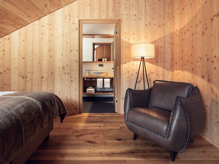 Una habitación con paneles de madera, una cama, un sillón de cuero y un baño visible a través de una puerta abierta.