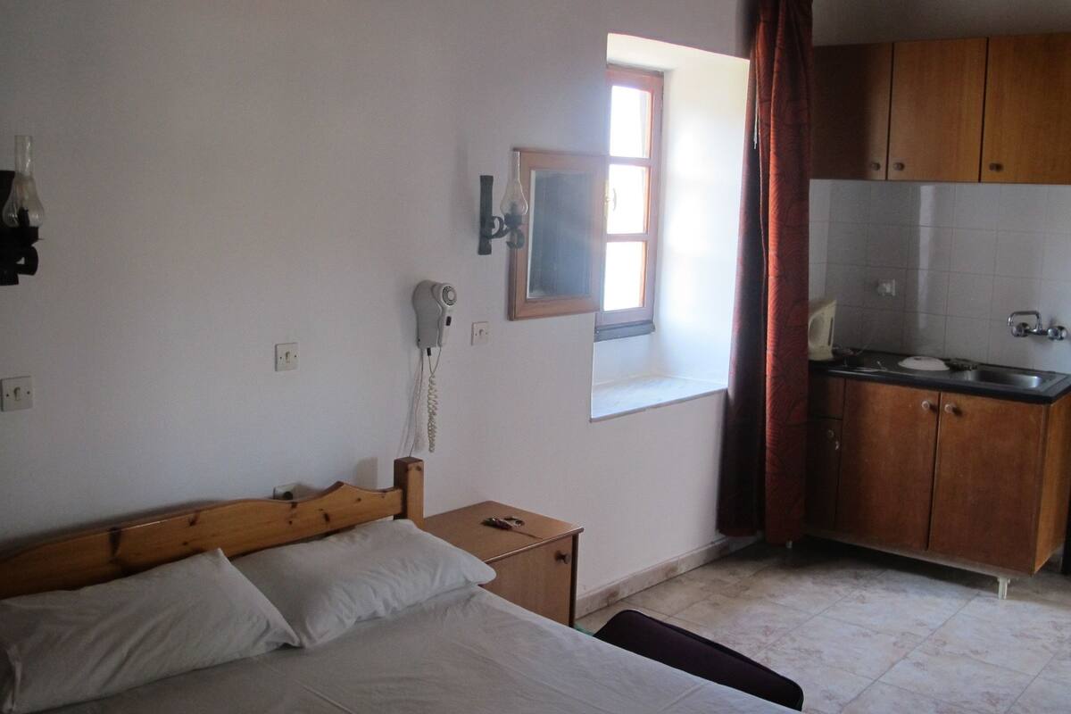 A bedroom with a wooden bed, a nightstand, a hairdryer, a window, and a kitchenette with wooden cabinets and a sink.