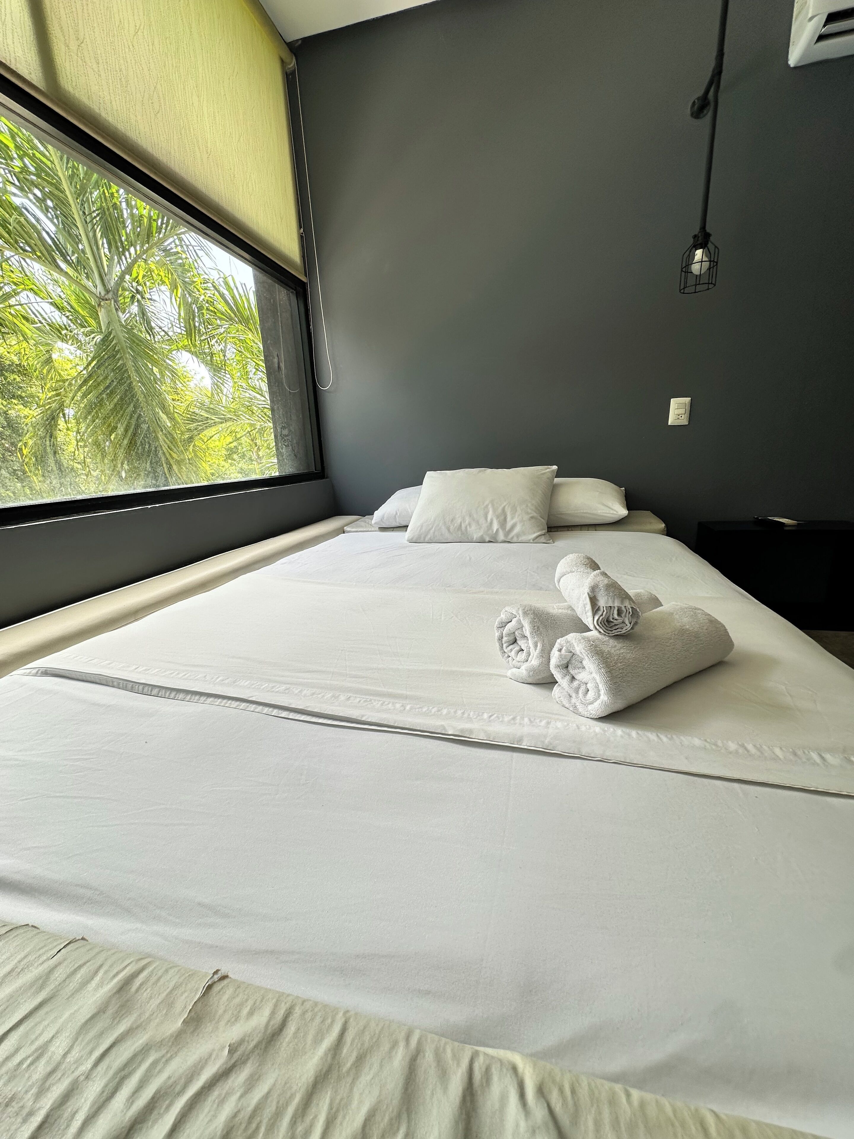 Una cama bien hecha con sábanas blancas y toallas enrolladas, una pared oscura y una ventana con vistas a palmeras.