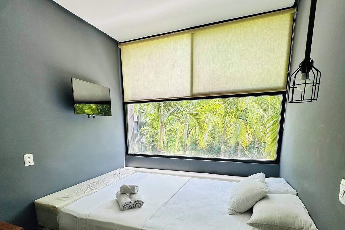 Ein modernes Schlafzimmer mit großem Fenster, einem Bett mit weißer Bettwäsche, einem kleinen Tisch und einem Nachttisch mit Lampe.