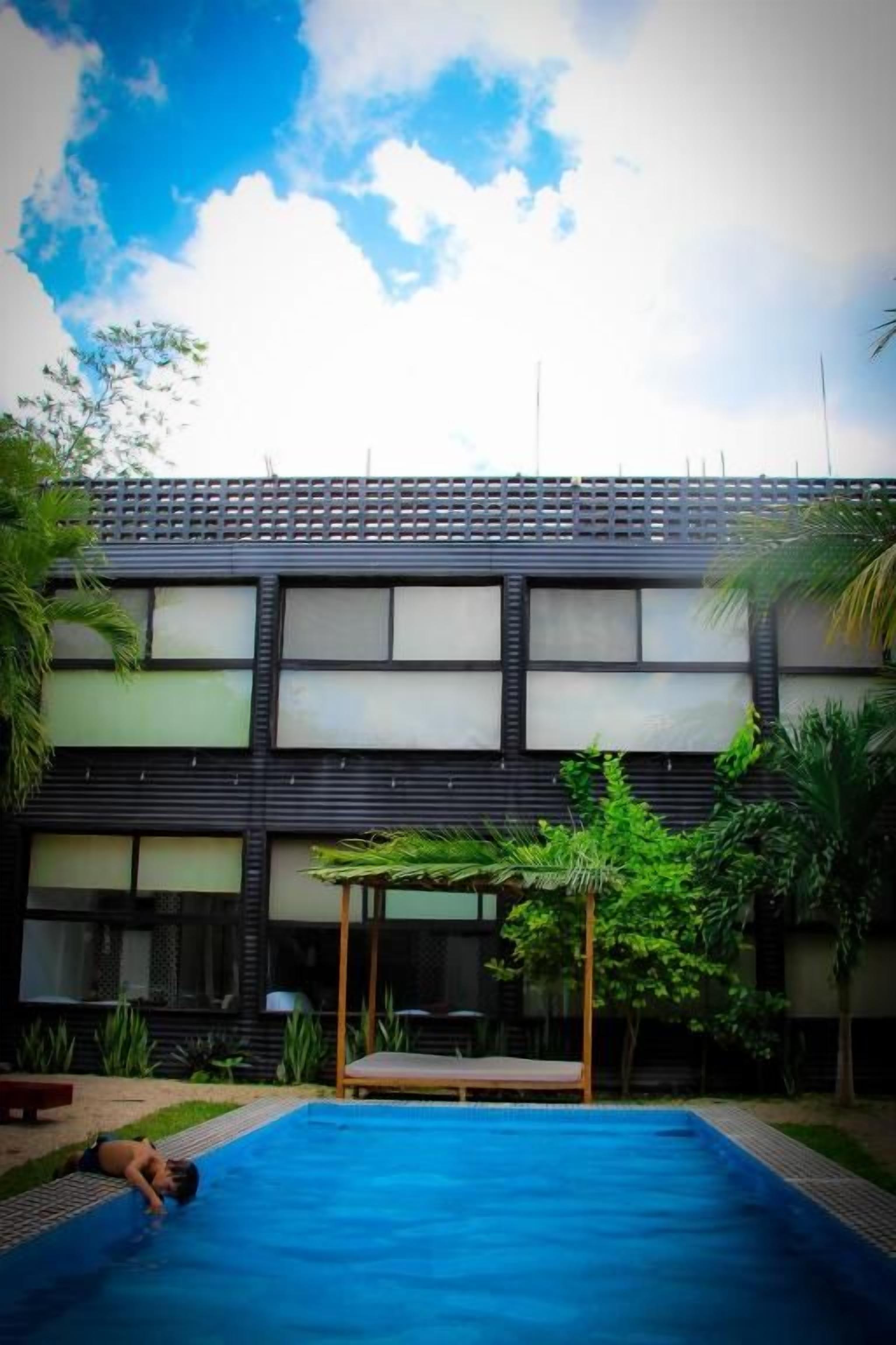 Una piscina con una persona recostada en el borde, rodeada de follaje y un edificio con amplios ventanales.