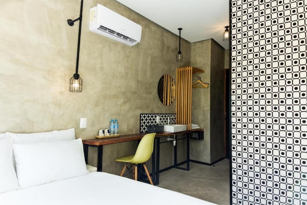 Une chambre d’hôtel comprenant un lit, un bureau avec une chaise, un luminaire suspendu, un climatiseur fixé au mur et un papier peint à motifs.