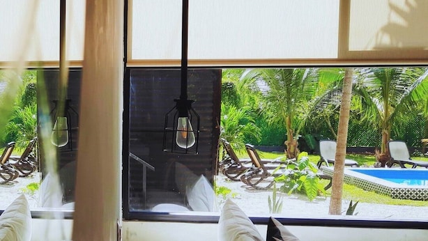 A room with a large window overlooking a pool and greenery. There is a wooden table with a remote control on it, and a white sofa with a pillow. - Bacalar 102 Hotel (Bacalar)