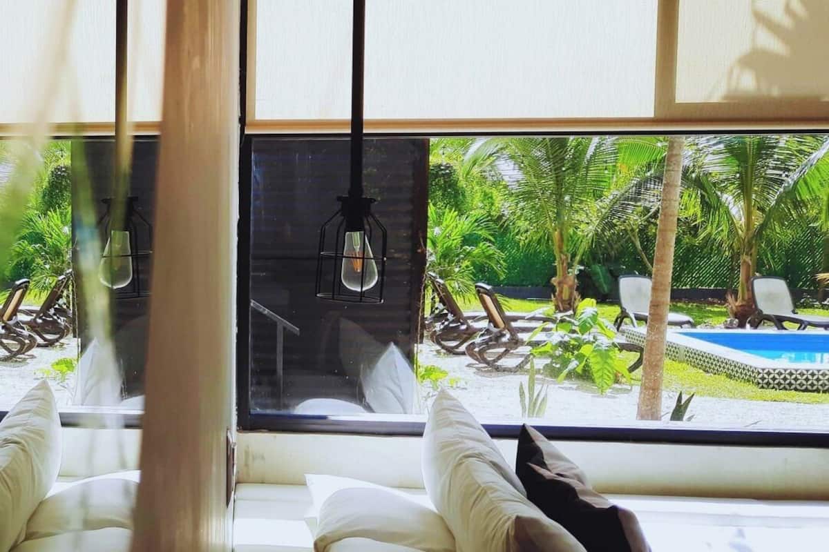 Ein Zimmer mit großem Fensterblick auf Pool und Grünflächen. Ein Holztisch mit Fernbedienung steht dort, sowie ein weißes Sofa mit Kissen.