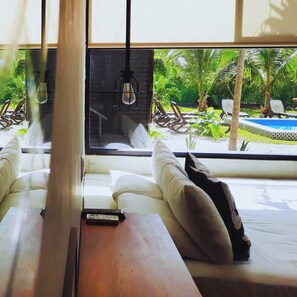 Ein Zimmer mit großem Fensterblick auf Pool und Grünflächen. Ein Holztisch mit Fernbedienung steht dort, sowie ein weißes Sofa mit Kissen.