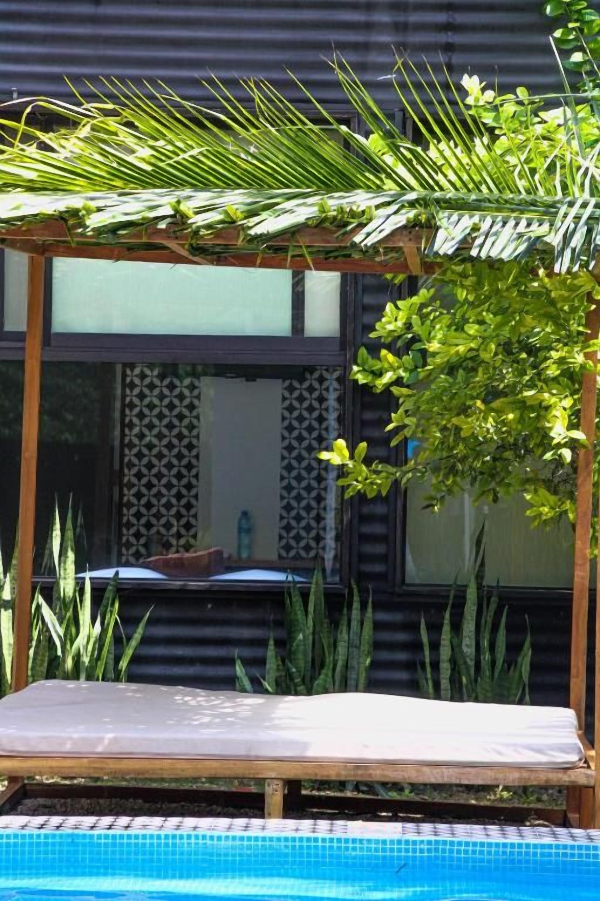 A pool area with a wooden pergola, a lounge chair, and tropical plants.