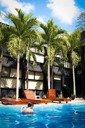 Piscina con palmeras y sillas de descanso, rodeada por un edificio con balcones.