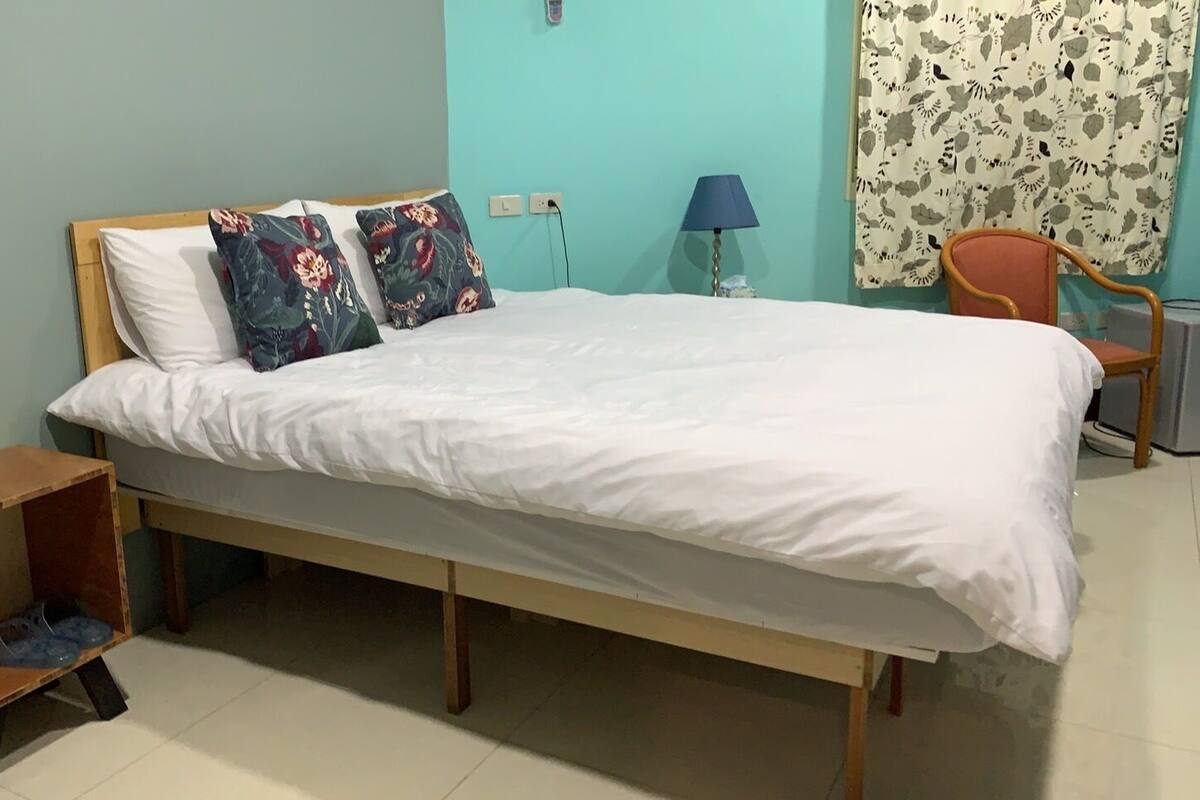 A single bed with white bedding, two floral pillows, a wooden headboard, a small wooden nightstand with a pair of slippers, a lamp, and a chair.