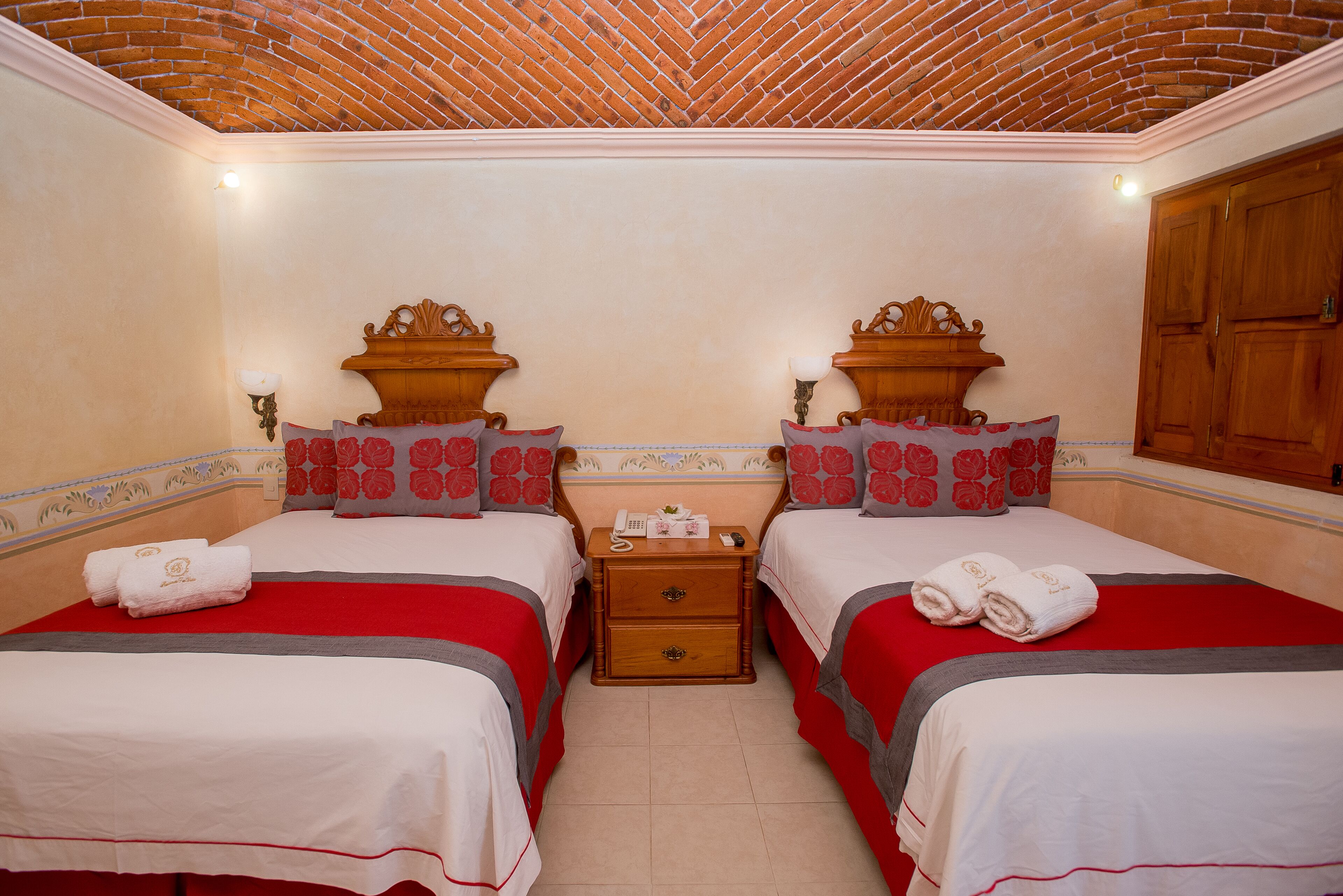 A hotel room with two beds, each with red and white bedding, a wooden nightstand with a telephone and a lamp, and a brick-patterned ceiling.