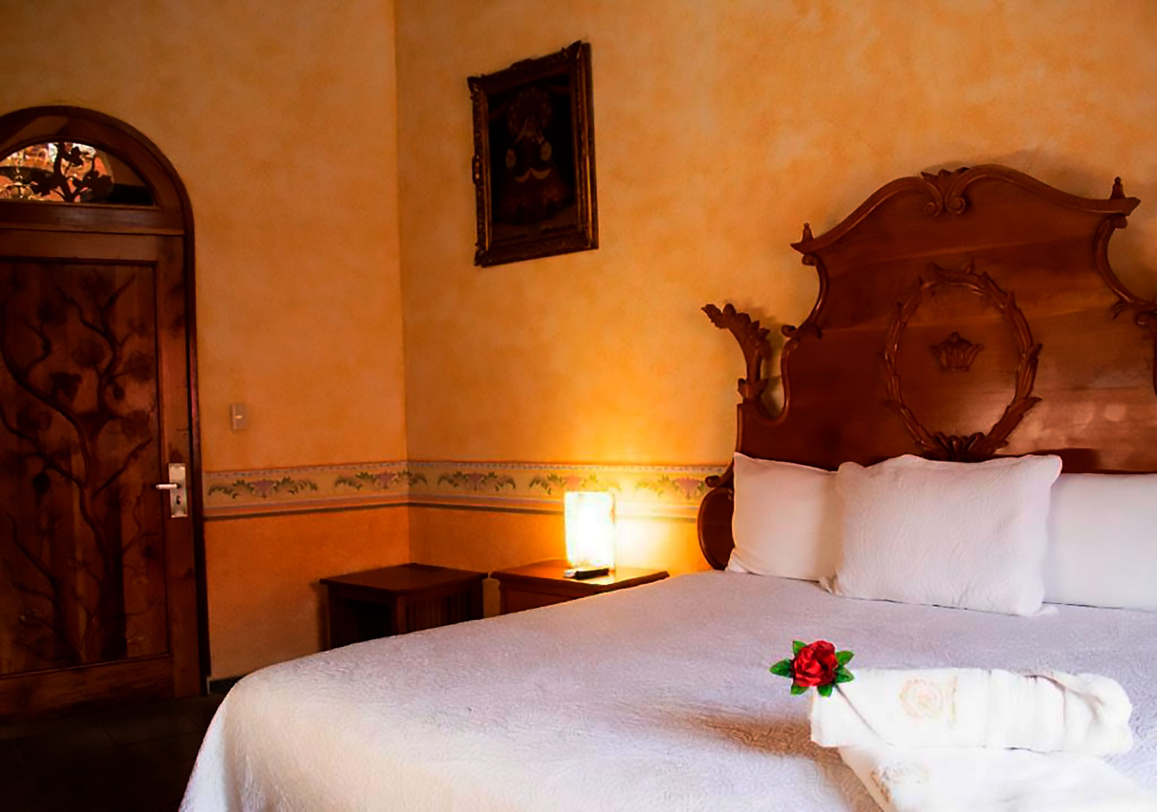 A hotel room with a wooden door, a bed with white linens, a bedside table with a lamp, and a framed picture on the wall.