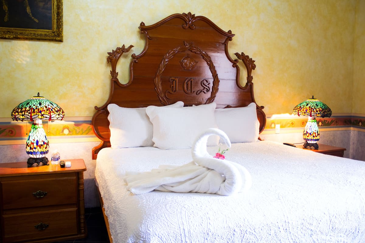 Een keurig opgemaakt bed met een handdoek in de vorm van een zwaan, een houten hoofdbord met een ornament, en twee lampen op nachtkastjes.