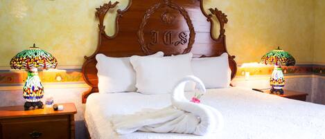 A neatly made bed with a swan-shaped towel, a wooden headboard with a crest, and two lamps on nightstands.