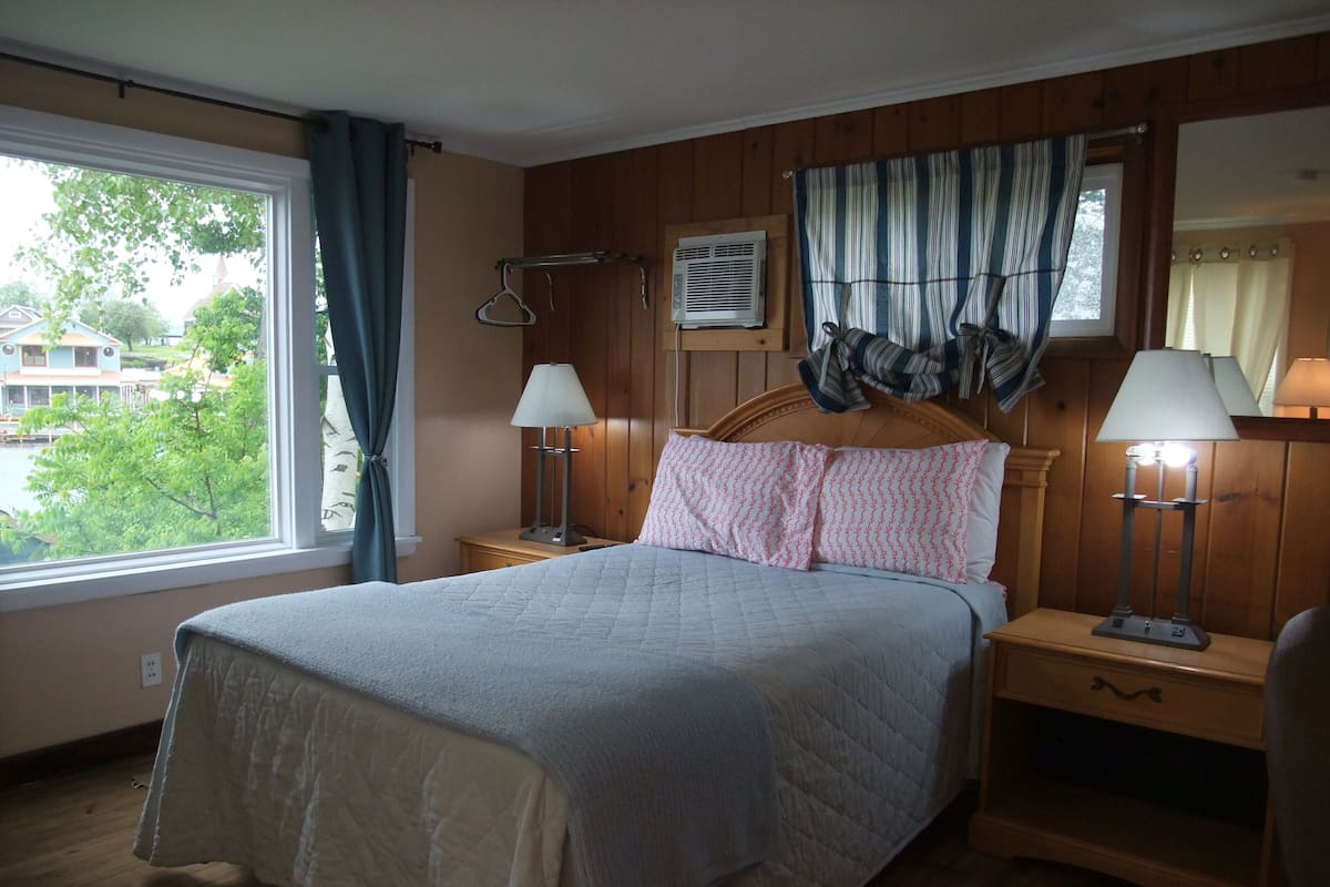 Een slaapkamer met een bed, twee nachtkastjes met lampen, een raam met gordijnen en uitzicht op huizen.