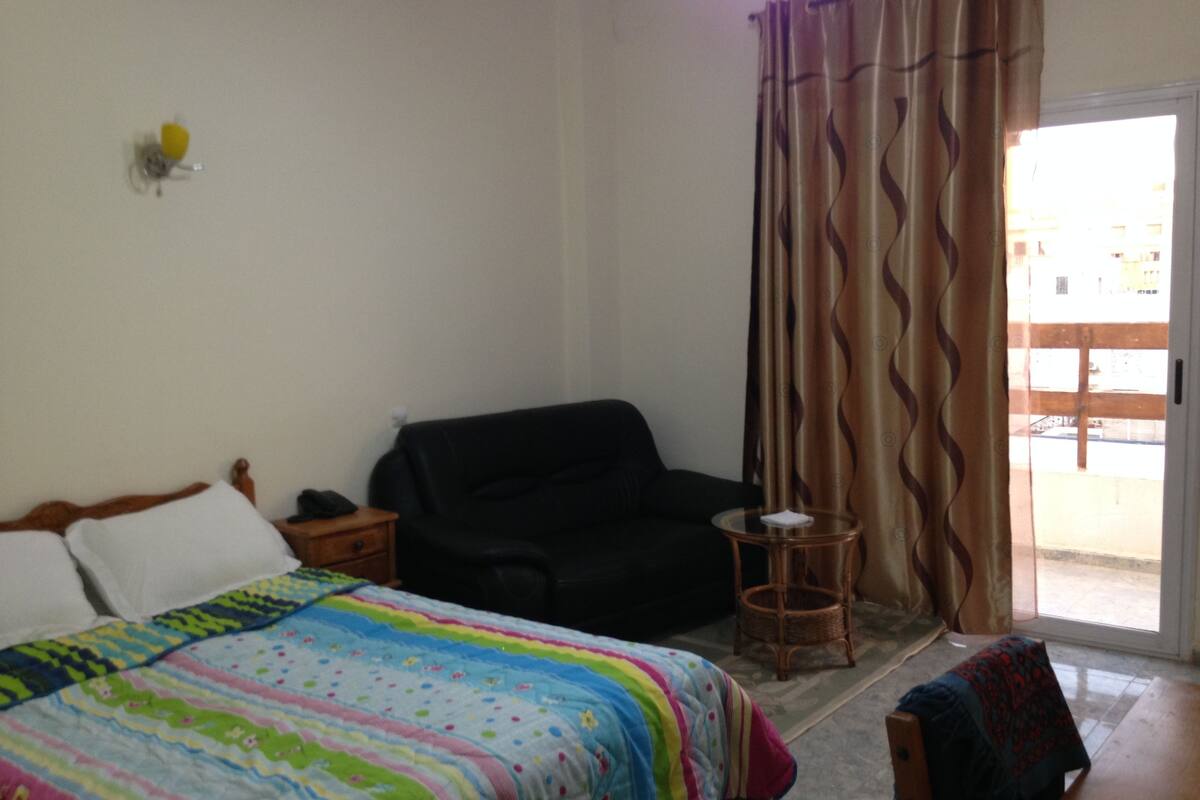 Un dormitorio con cama, una silla negra, una mesita y una ventana con cortinas.