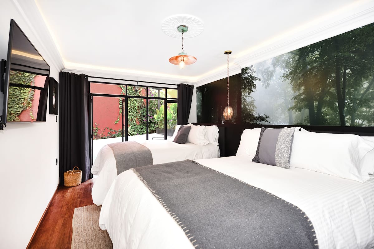 Quarto de hotel com duas camas, uma janela grande com vista para uma floresta e um lustre.