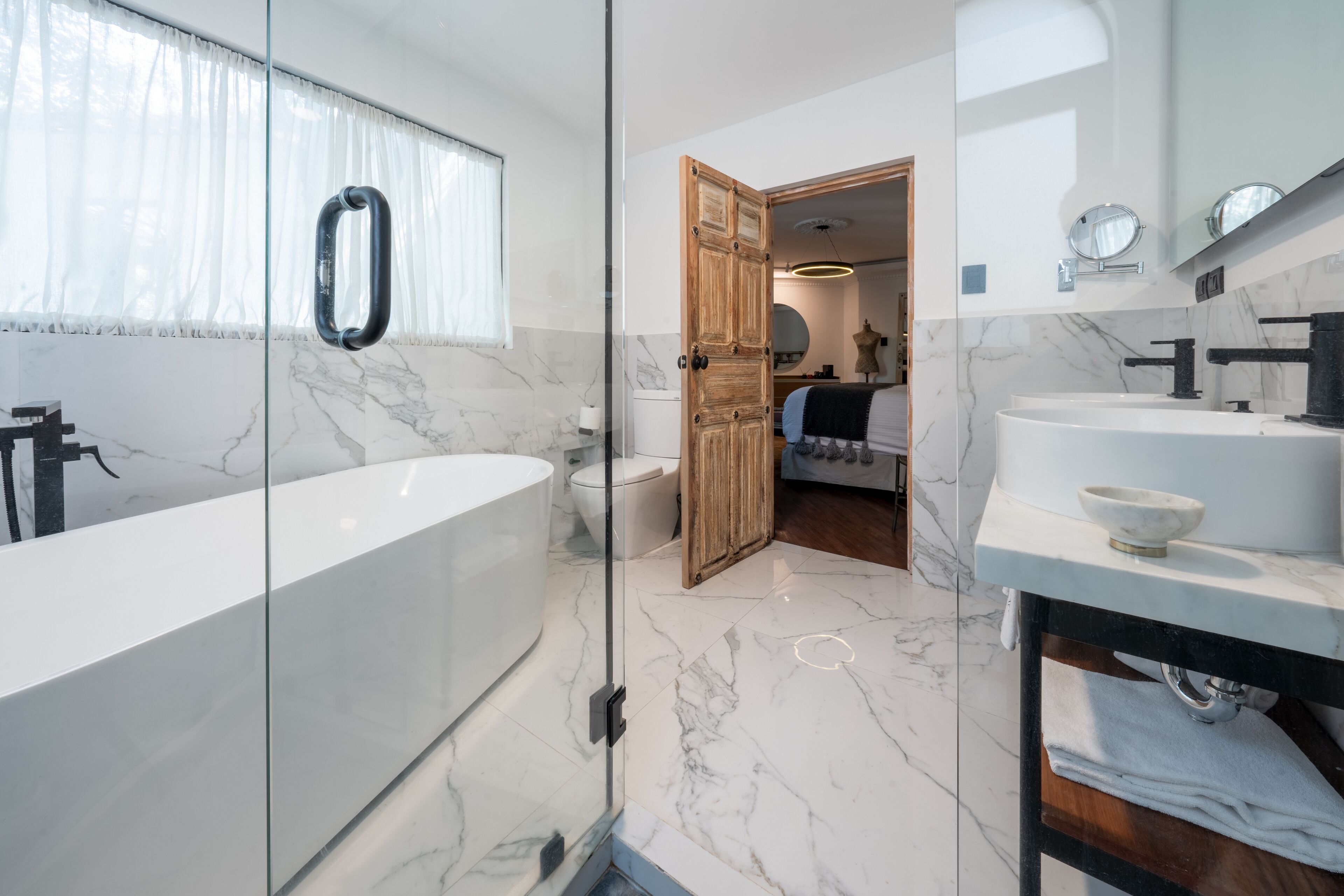 A modern bathroom with a glass-enclosed shower, a marble-tiled floor, a white sink, and a wooden door leading to another room.