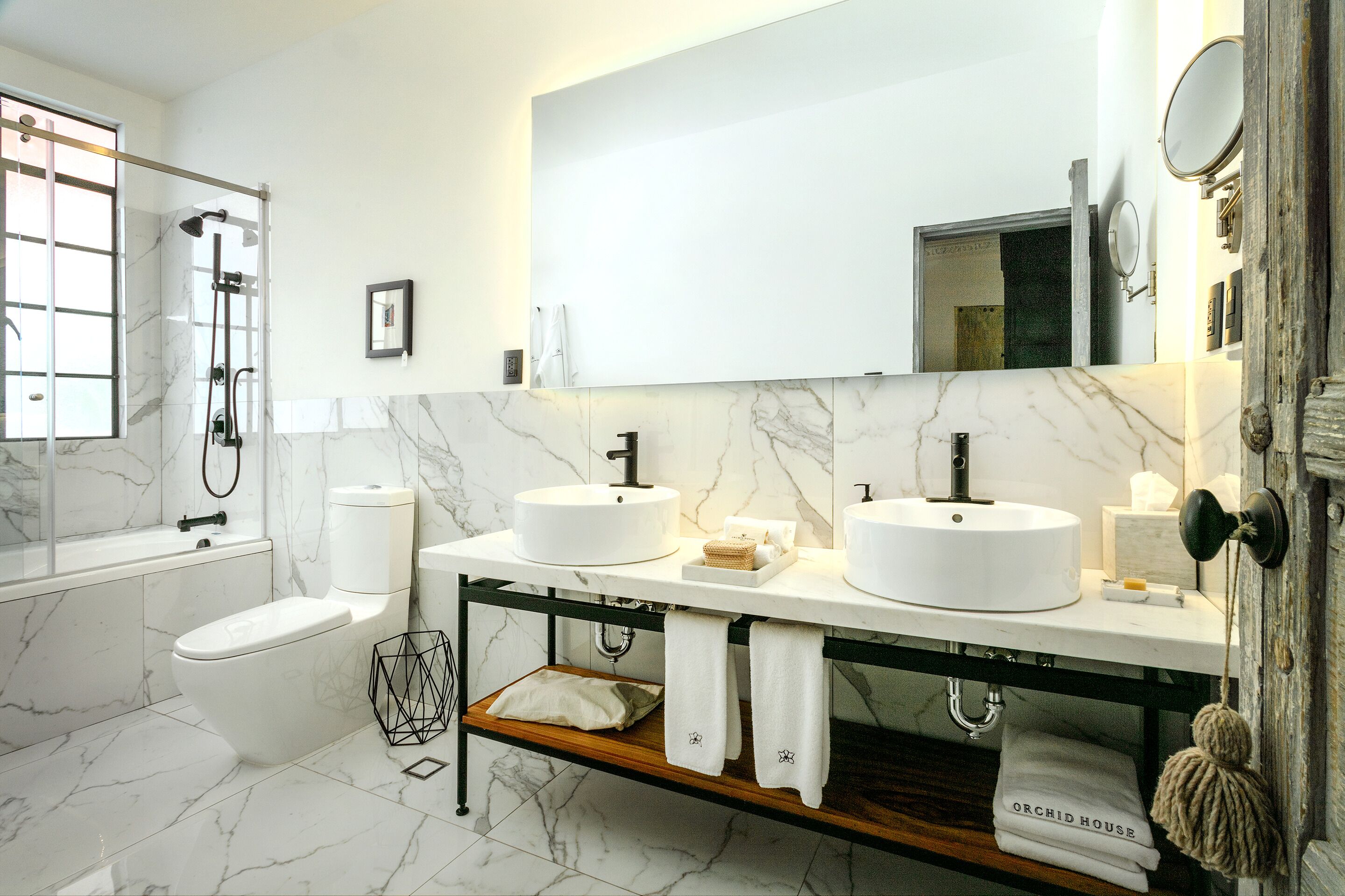 Un baño moderno con dos lavamanos, un inodoro y una ducha. Hay un estante de madera debajo de los lavamanos y un perchero con toallas.