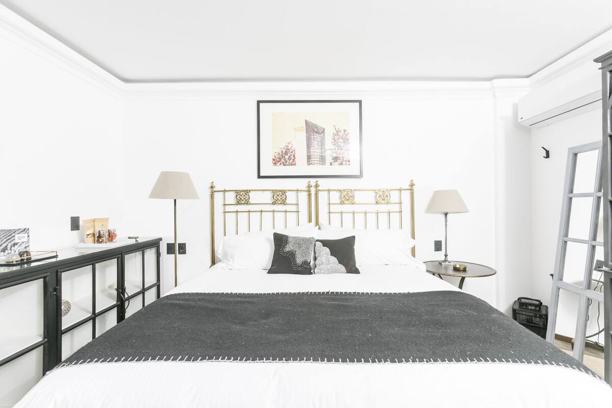 Quarto bem arrumado com cama, mesas de cabeceira, abajures e um quadro na parede.