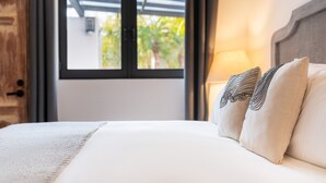Una cama bien hecha con un edredón blanco y dos almohadas decorativas, una puerta de madera y una ventana con vistas a la vegetación.