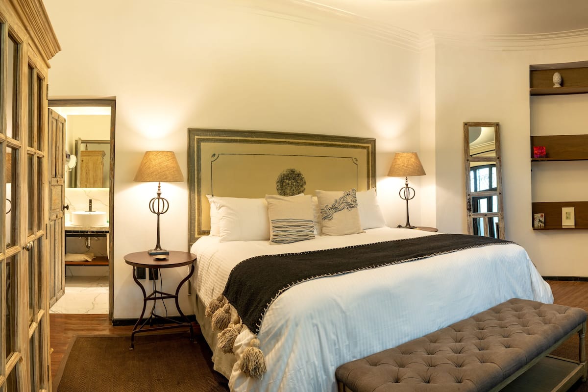 Quarto bem iluminado com uma cama grande, mesas de cabeceira, um banquinho, televisão e uma estante com objetos decorativos.