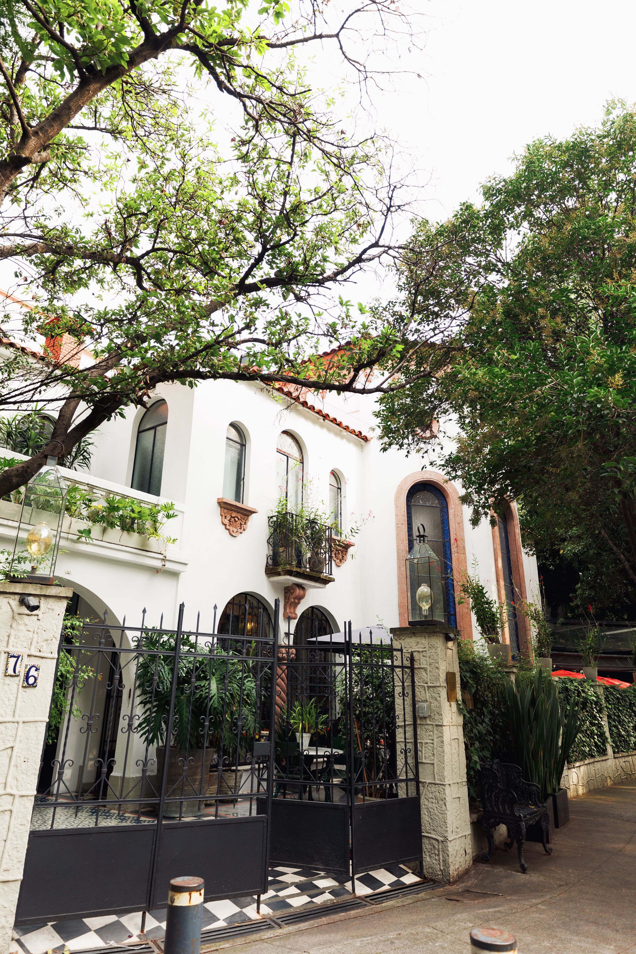Un edificio blanco con ventanas arqueadas y balcones, rodeado por una valla de hierro negro y macetas con plantas.