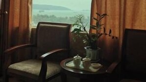 Une pièce avec vue sur un paysage lointain, visible à travers une fenêtre, comprenant une chaise, une petite table avec un plateau et une plante en pot.