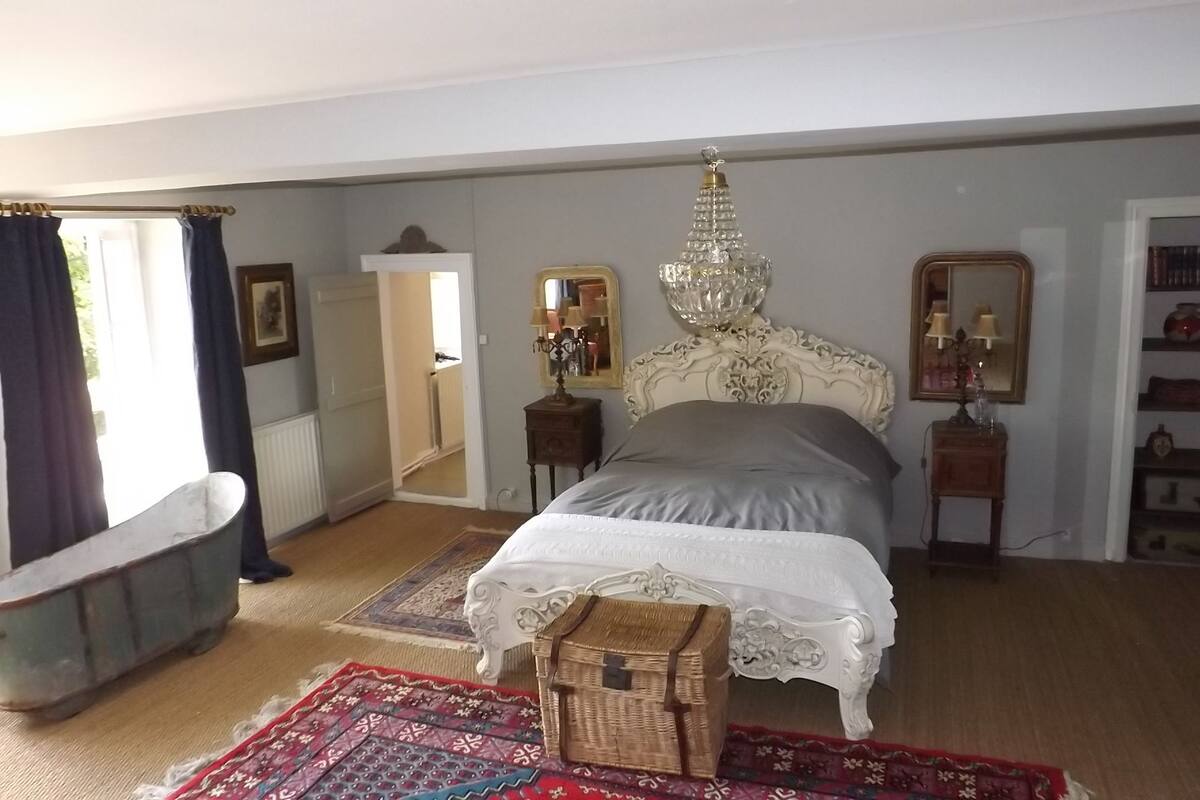 Een slaapkamer met een groot bed, een kroonluchter, een dressoir, een spiegel en een boekenplank.