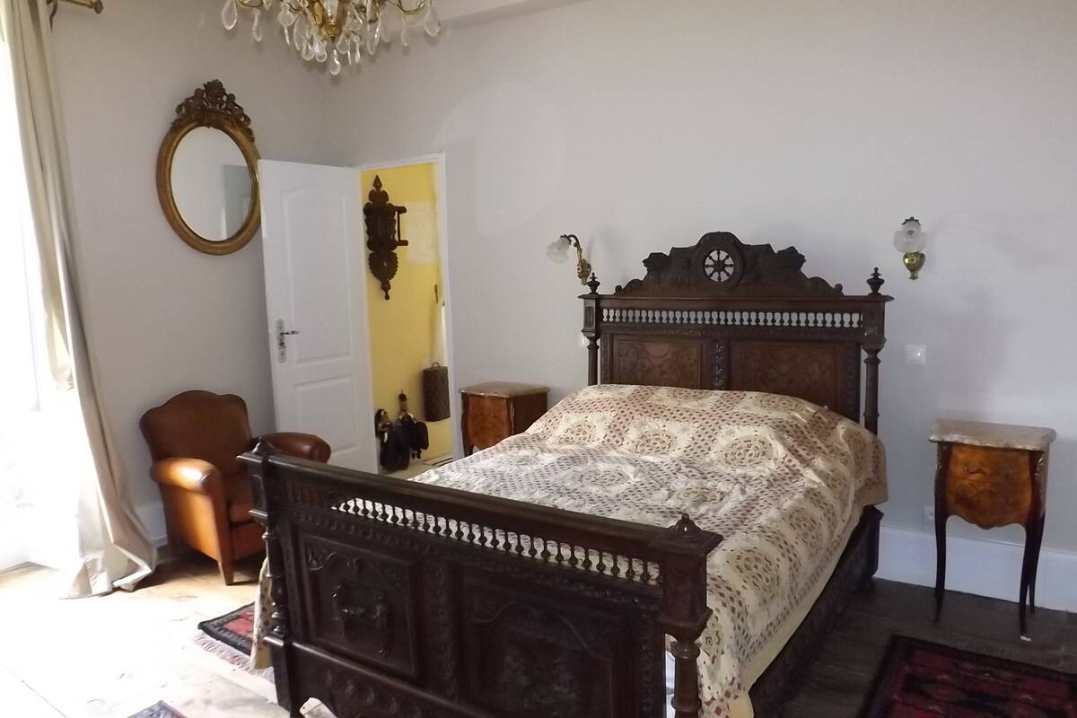 Een slaapkamer met een houten bed, een stoel, een tafeltje, een spiegel en een deur.